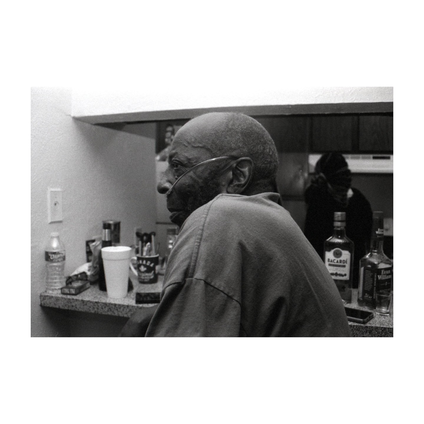 A man with an oxygen tube in his nose sits at a counter with bottles of liquor and other items.