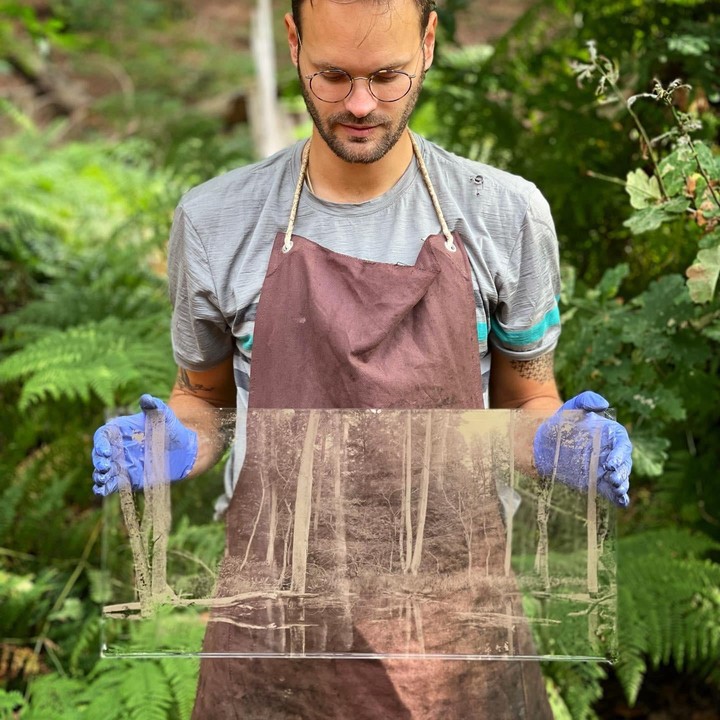 A man in an apron and blue gloves holds a large glass plate with a sepia-toned forest image on it.