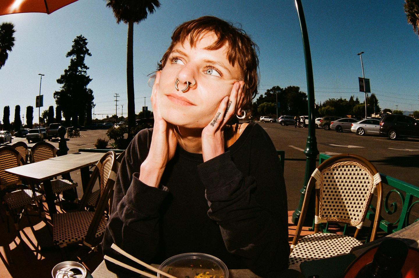 A woman with a nose ring sits at an outdoor cafe table with her hands on her face, looking up and to the side.