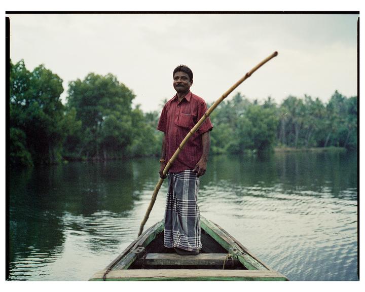 A man stands in a boat on a body of water, holding a long pole, with trees in the background.