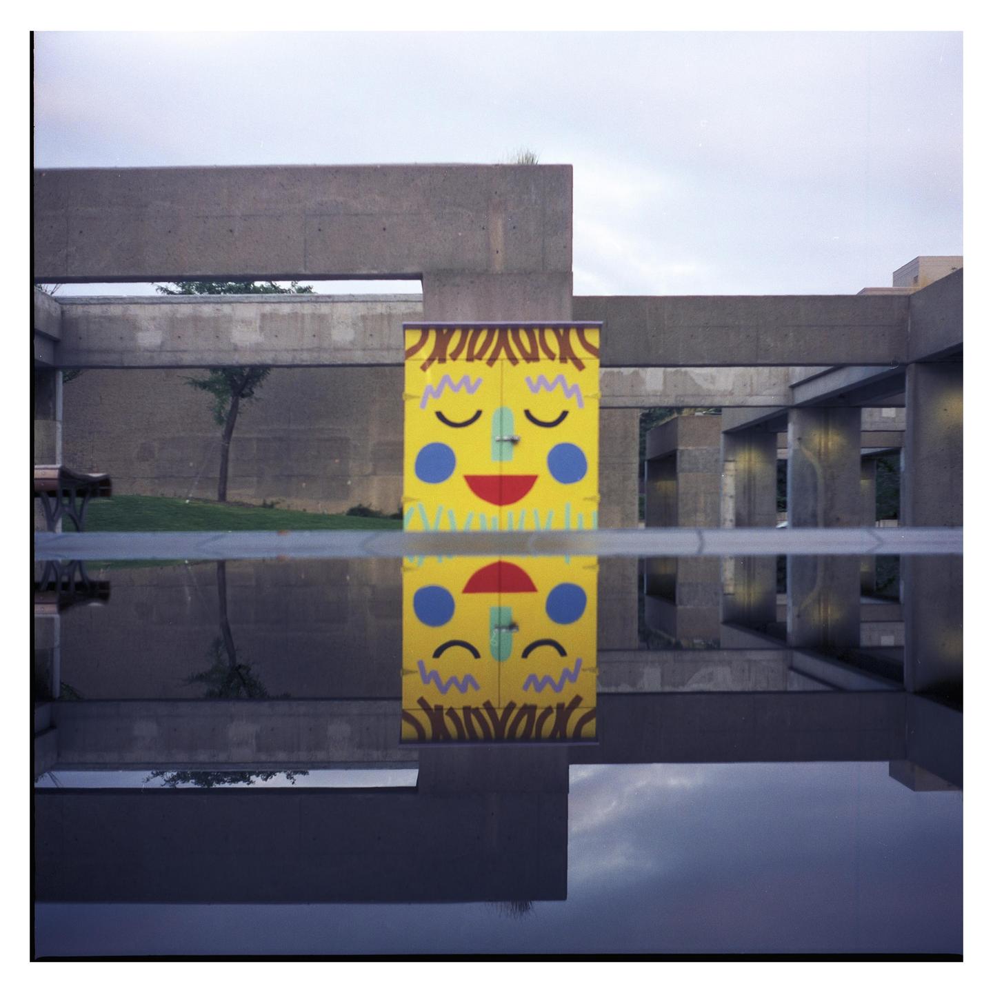 A yellow box with a smiley face is reflected in a pool of water in front of a concrete building.