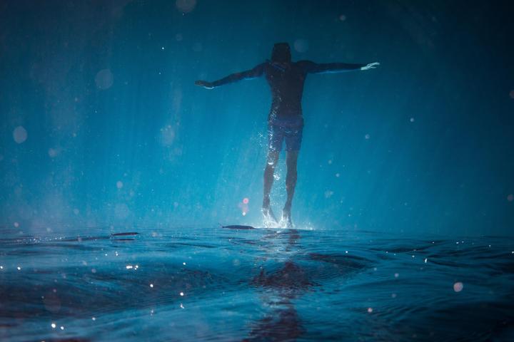 A man in a wetsuit is suspended in blue water with light particles and ripples on the surface.