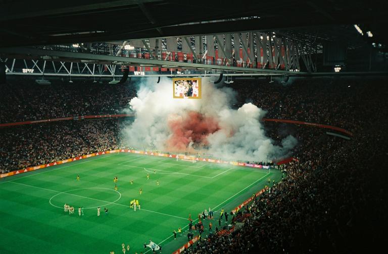 A soccer stadium is filled with spectators as smoke billows from the field during a game.