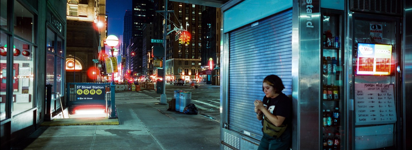 A woman stands next to a closed newsstand on a city street at night, with a subway station entrance and tall buildings in the background.