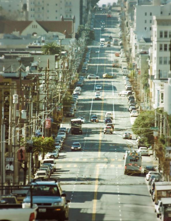 A long, steep street with cars parked on both sides and driving in both directions is lined with buildings and utility poles.