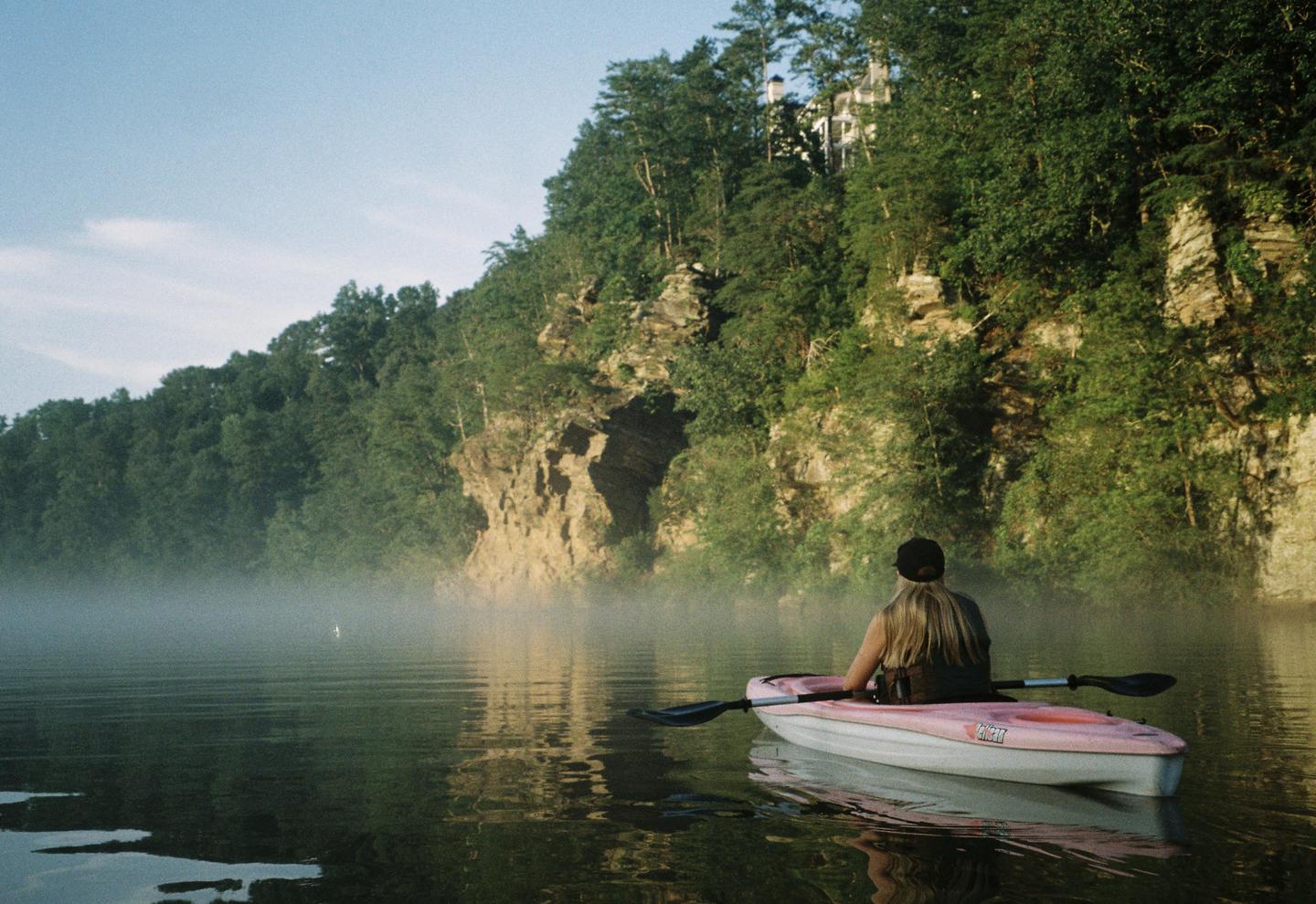 A woman in a pink kayak paddles on a misty lake surrounded by a rocky, forested shore.