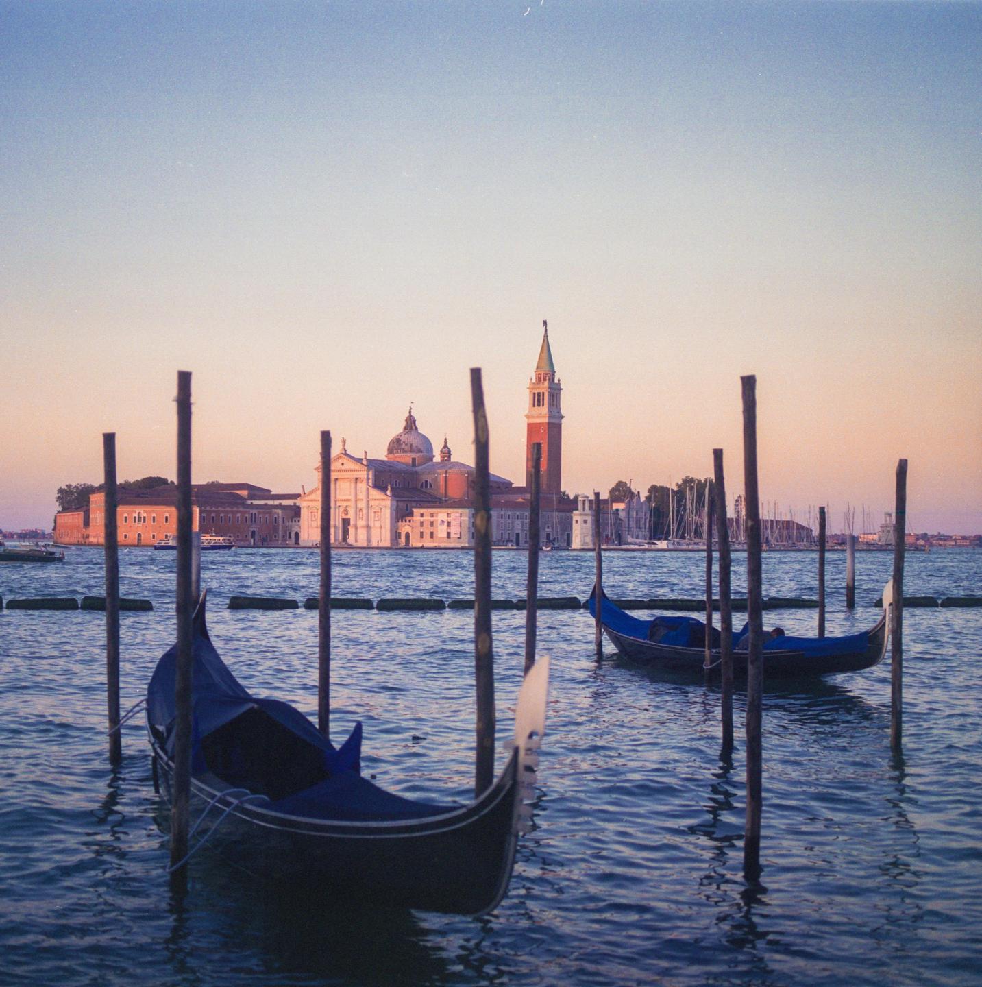 Two gondolas are moored in the water with a city and bell tower in the background.