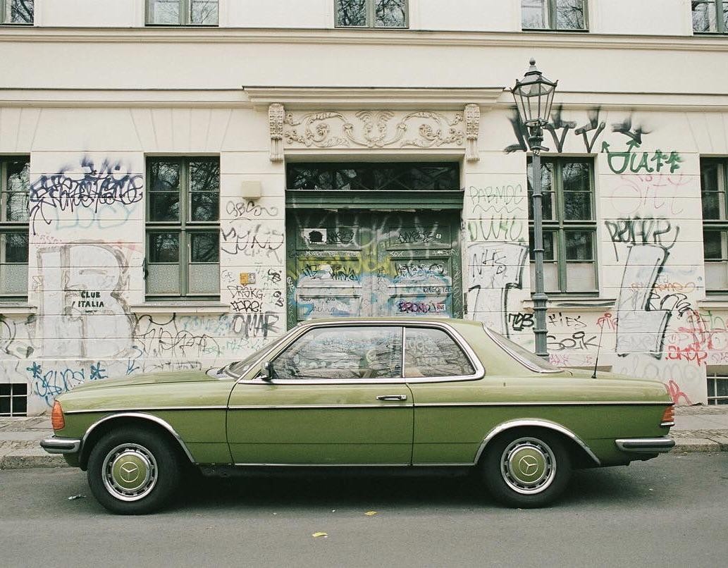 A green car is parked on a street in front of a building covered in graffiti.
