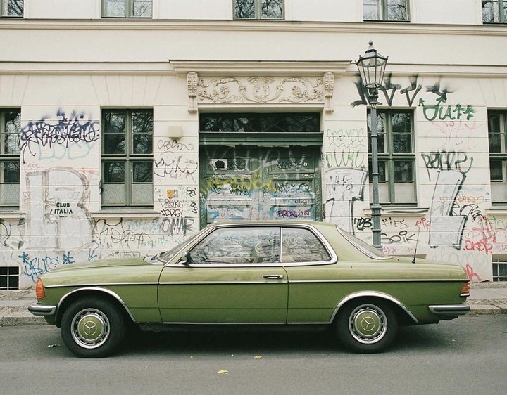 A green car is parked on a street in front of a building covered in graffiti.