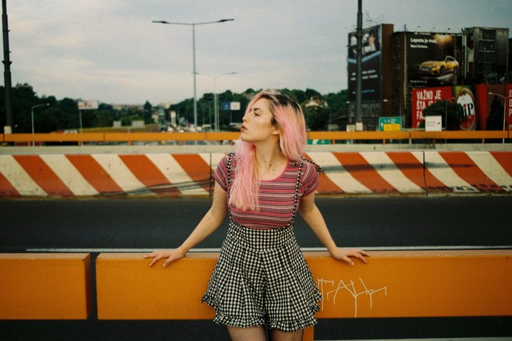 A woman with pink hair stands on a bridge next to a road barrier with orange and white stripes.