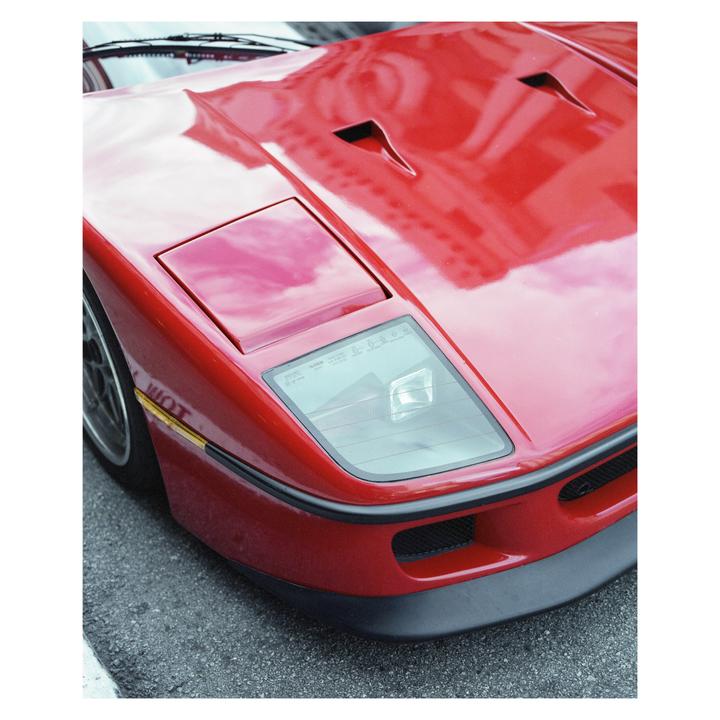 A red sports car is parked on a street, with reflections of clouds and buildings visible on its shiny surface.