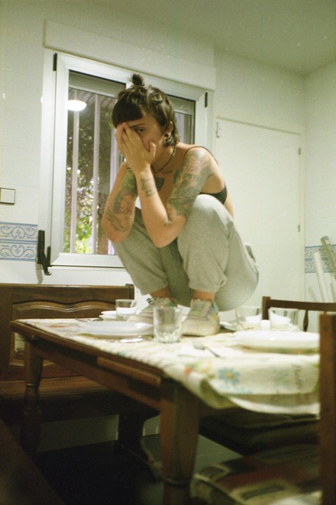 A woman crouches on a dining table, covering her face with her hands.
