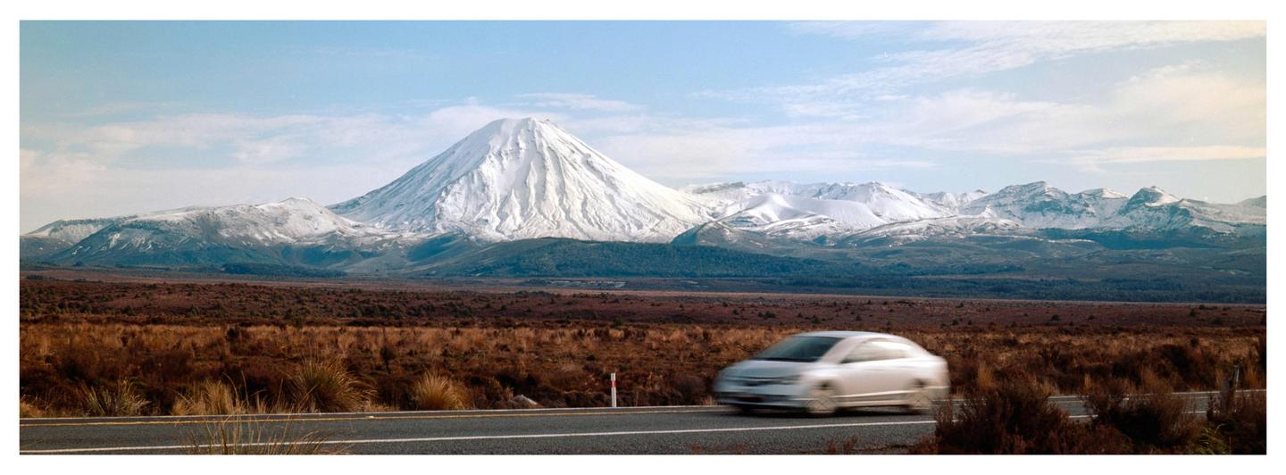 A white car drives on a road with a snow-capped mountain in the background.