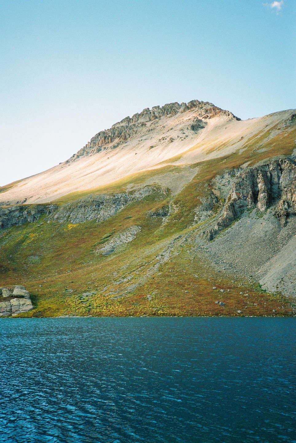 A large mountain with patches of green and brown foliage rises from the edge of a deep blue lake under a clear sky.