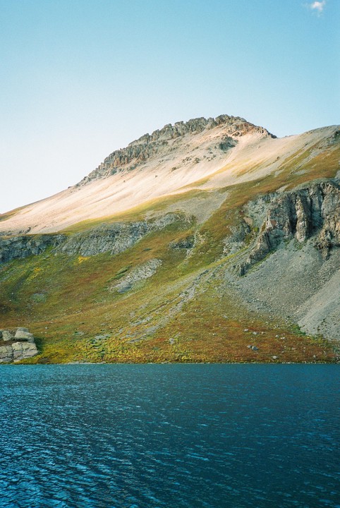 A large mountain with patches of green and brown foliage rises from the edge of a deep blue lake under a clear sky.