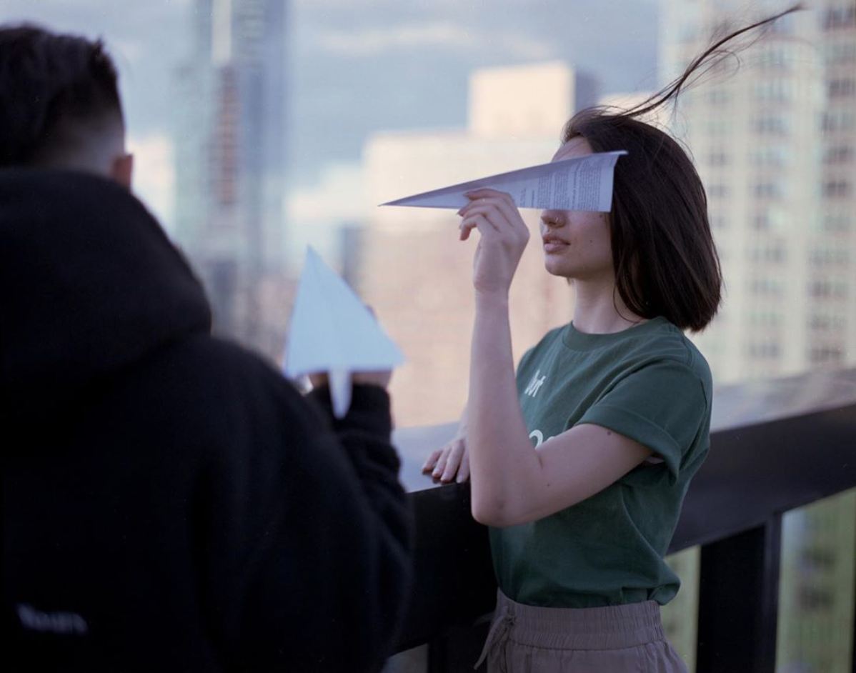A woman holds a paper airplane over her eyes while a man holds another paper airplane in the foreground.