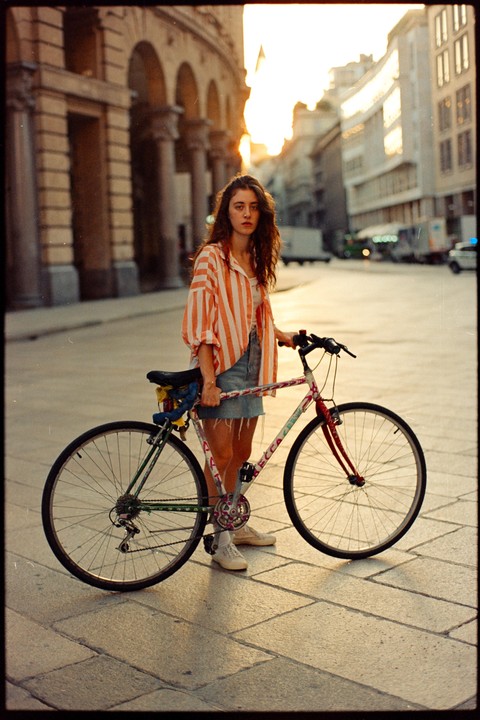 A woman stands next to her bicycle on a paved street with buildings in the background at dawn.