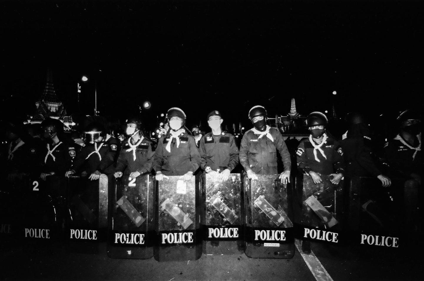 A line of police officers in uniform and helmets stand behind riot shields that say "POLICE" at night.