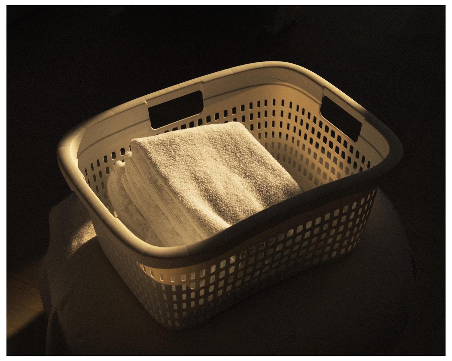 A white laundry basket filled with folded towels sits in a beam of light.