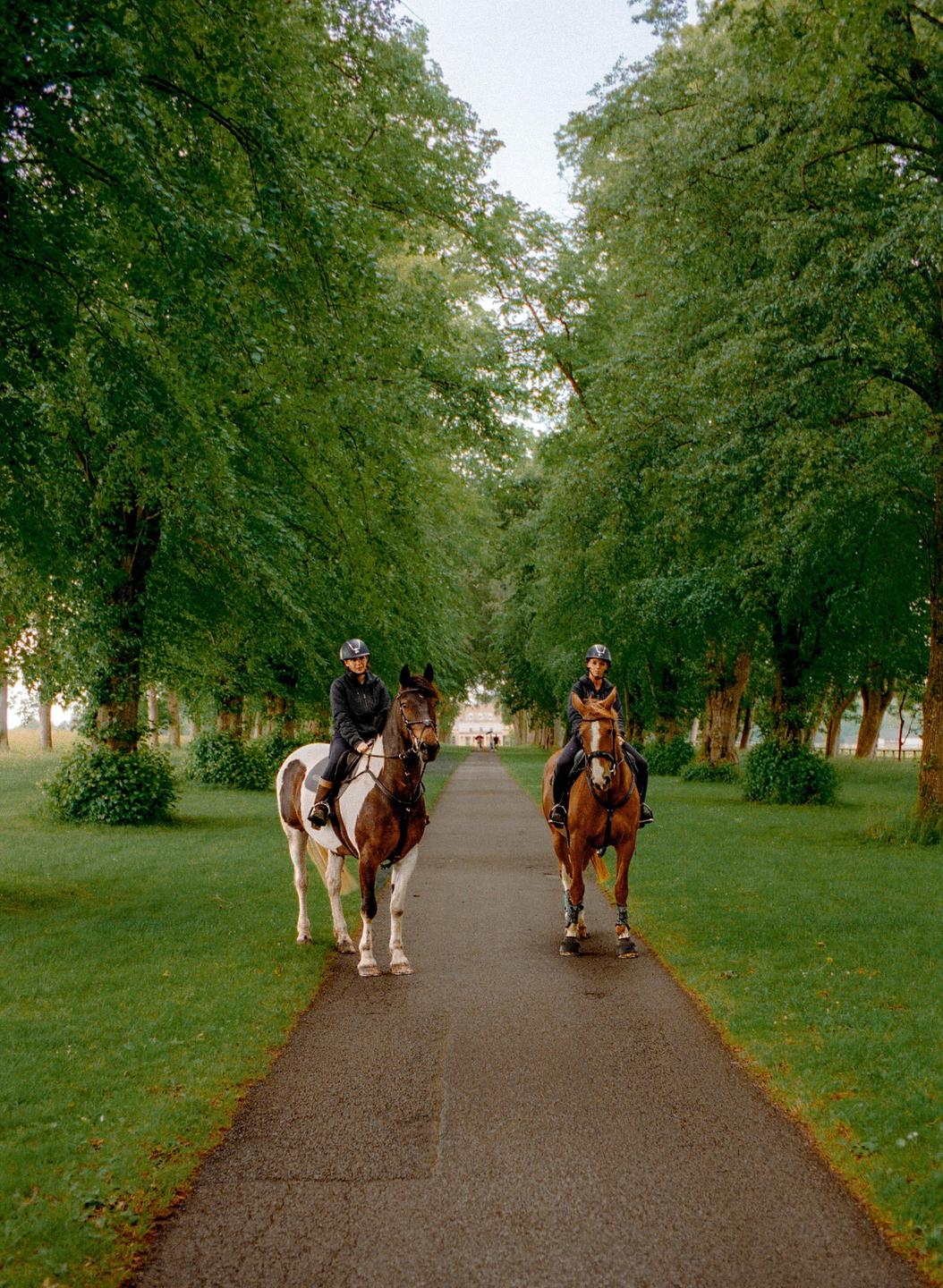Two people on horseback ride down a tree-lined path with green grass on either side.