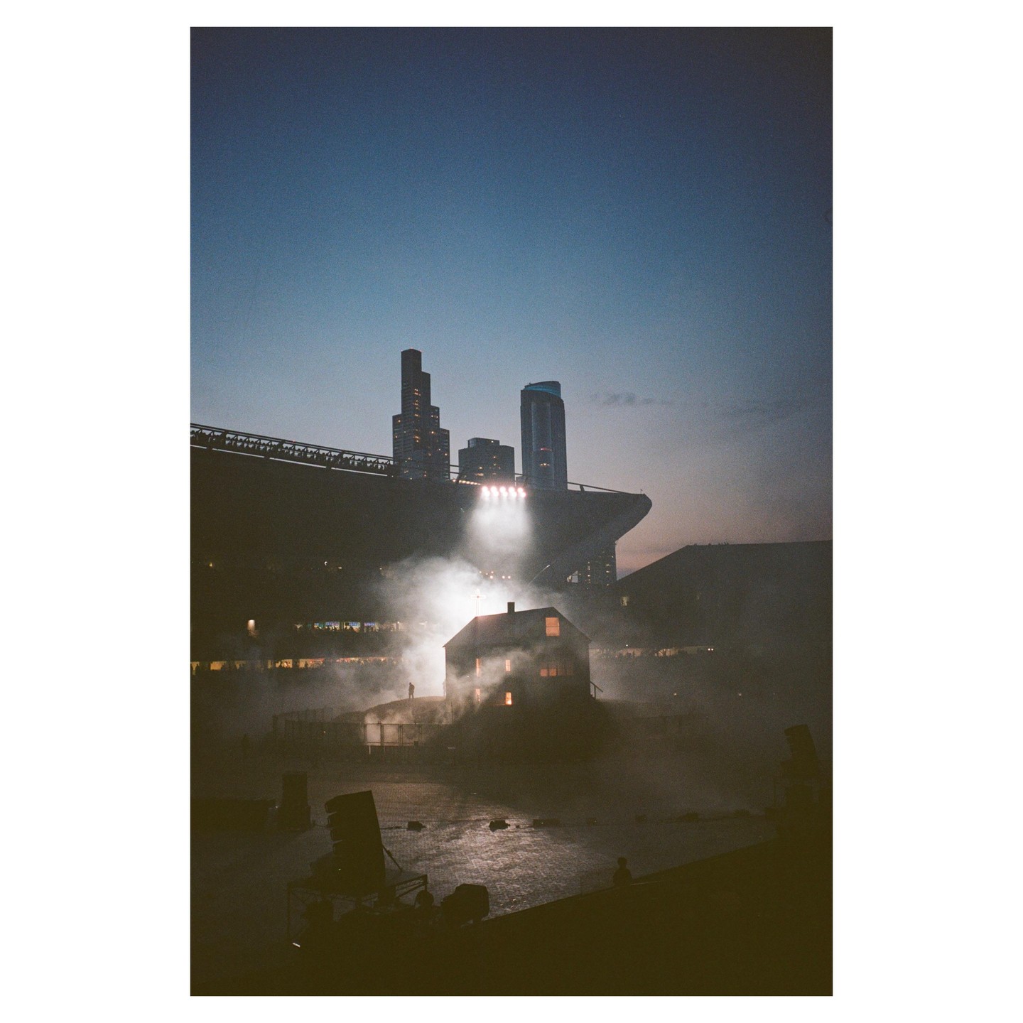 A small house is illuminated by spotlights and surrounded by smoke in a stadium at dusk, with city buildings visible in the background.