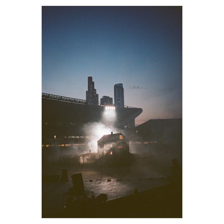 A small house is illuminated by spotlights and surrounded by smoke in a stadium at dusk, with city buildings visible in the background.