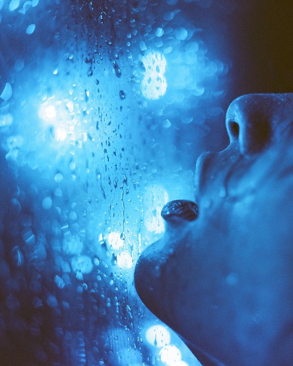 A person's face is visible in profile next to a window covered in raindrops and blue light.