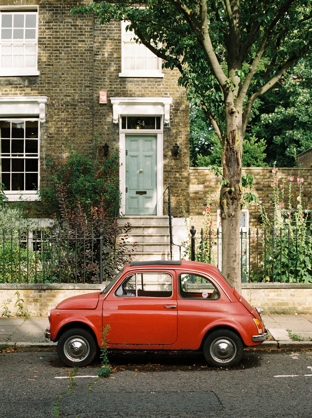 A red Fiat 500 is parked on a street in front of a brick house with a light blue door.