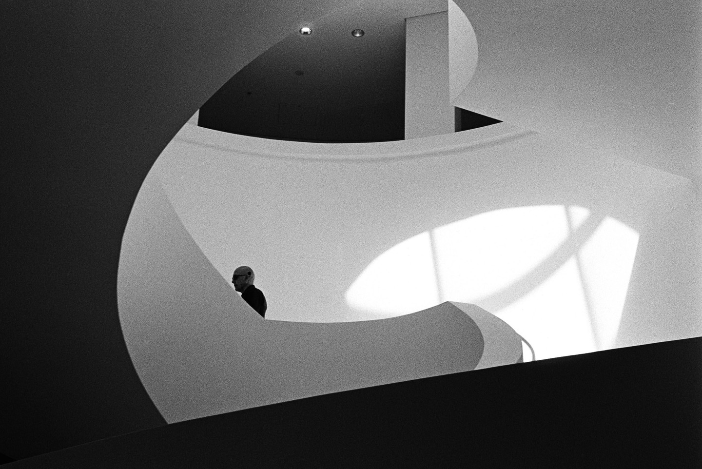 A man stands on a curved staircase with light and shadow patterns on the wall.