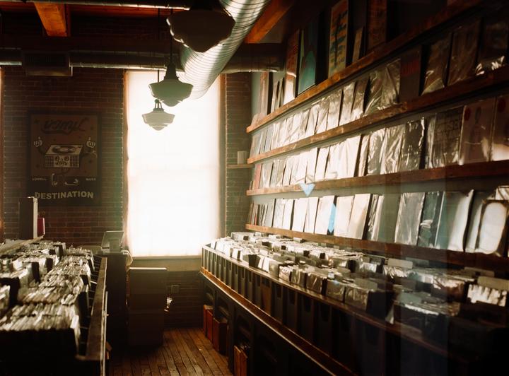A record store with brick walls and wooden floors is filled with rows of vinyl records and a large window at the back.