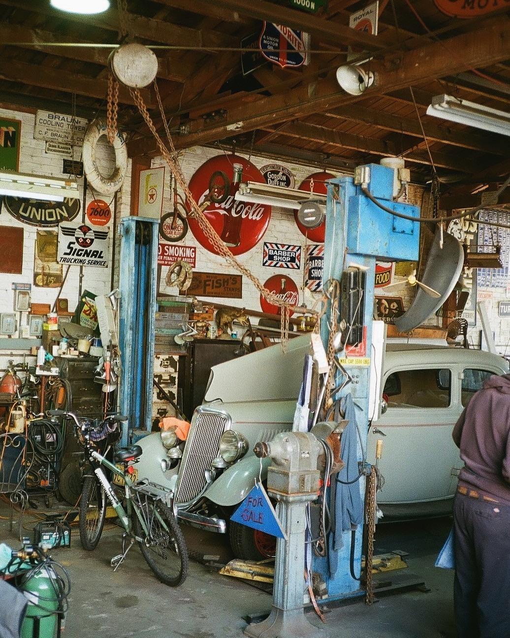 A vintage car is parked inside a cluttered garage filled with various signs and equipment.