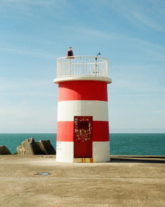 A red and white striped lighthouse stands on a concrete pier next to the ocean under a clear sky.