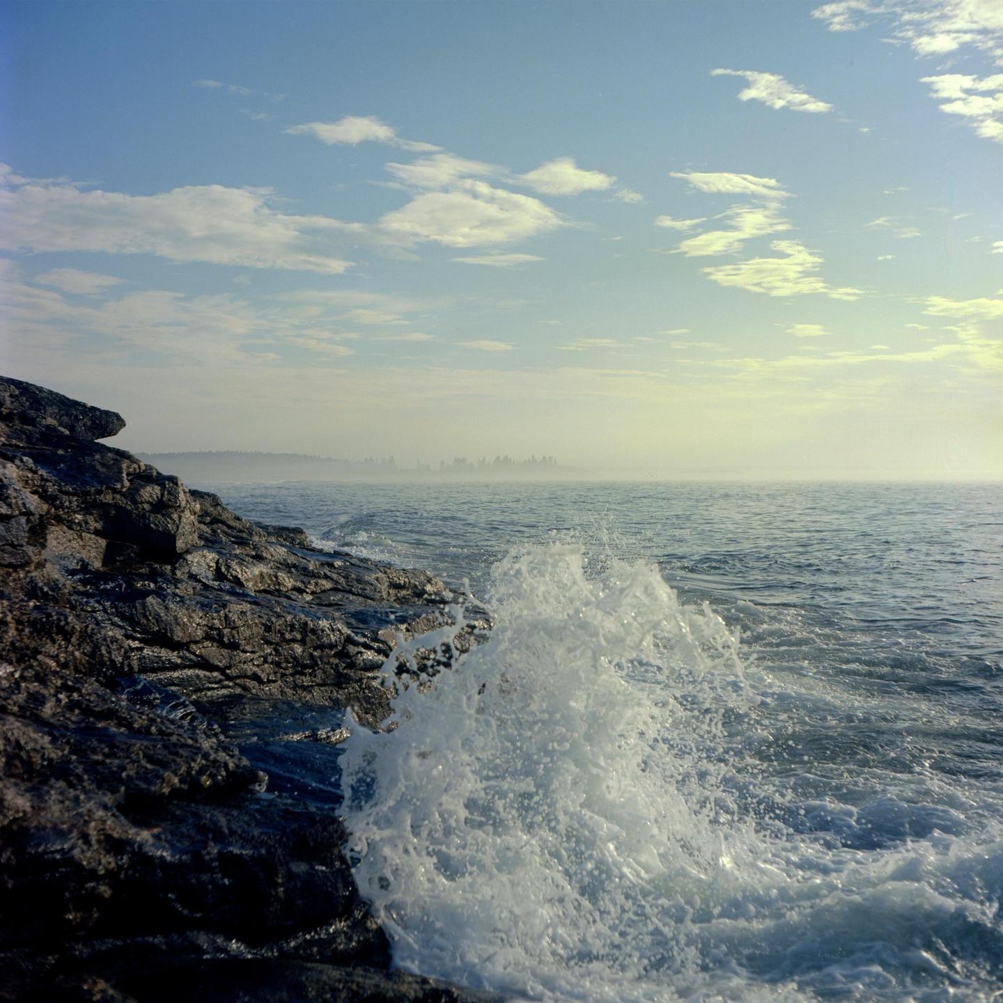 A wave crashes against a rocky shore under a partly cloudy sky.