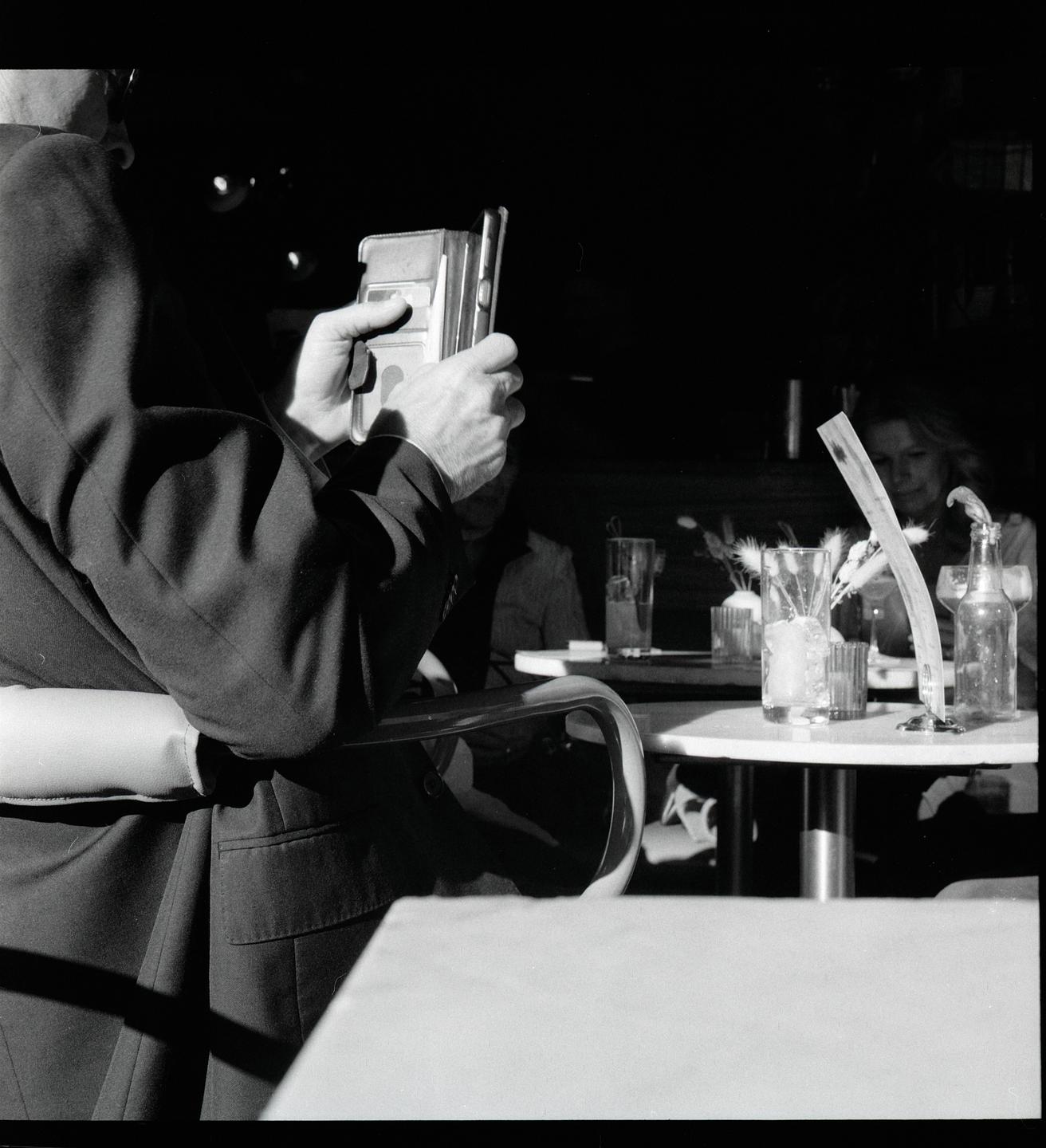 A man holds a phone at a table with drinks and a woman in the background.