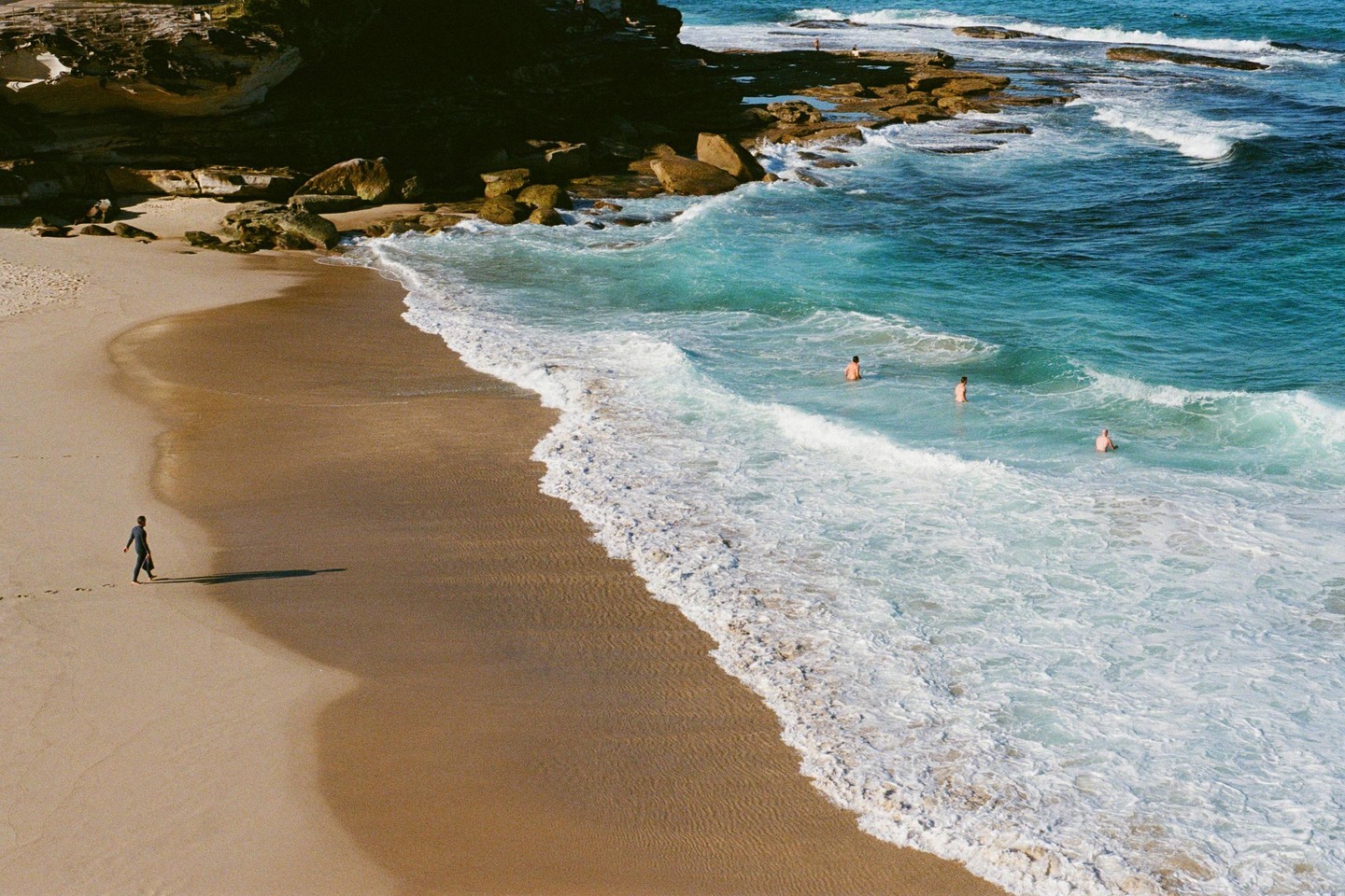A person walks along a sandy beach with waves crashing on the shore and three people swimming in the ocean.