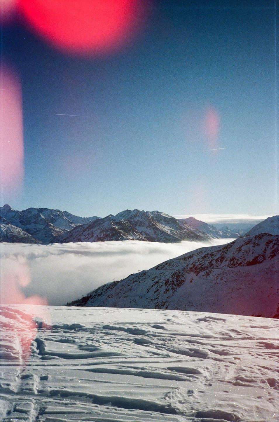 A snowy mountain landscape with a layer of clouds covering the valley and a bright red light leak in the upper left corner.