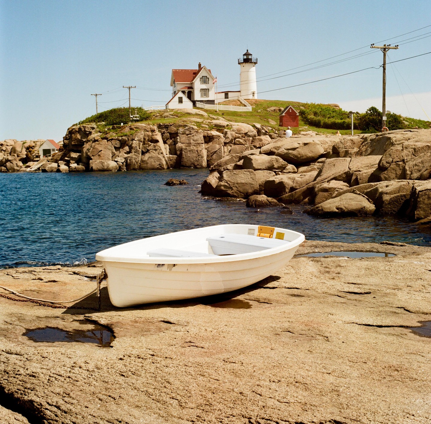 A white boat rests on a rocky shore with a lighthouse and house on a hill in the background.