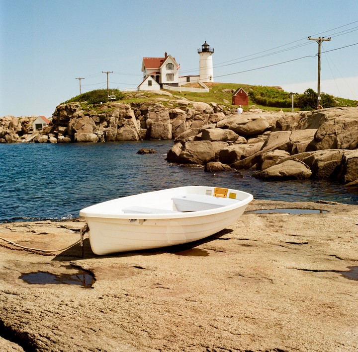 A white boat rests on a rocky shore with a lighthouse and house on a hill in the background.