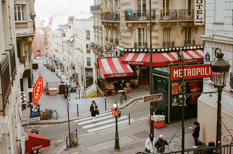 A woman crosses a street with a cafe and a metro station entrance on a hill in Paris.