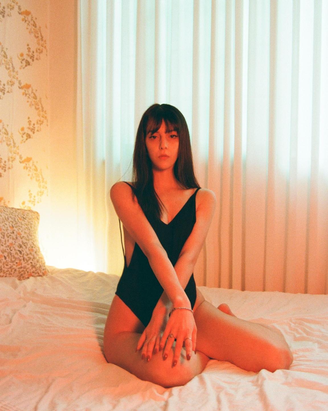 A woman in a black swimsuit sits on a bed with her arms crossed, looking directly at the viewer.