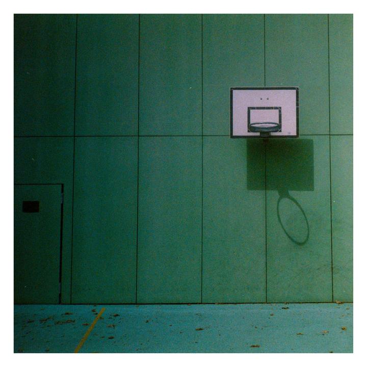 A basketball hoop casts a shadow on a green wall above a green court with a yellow line.