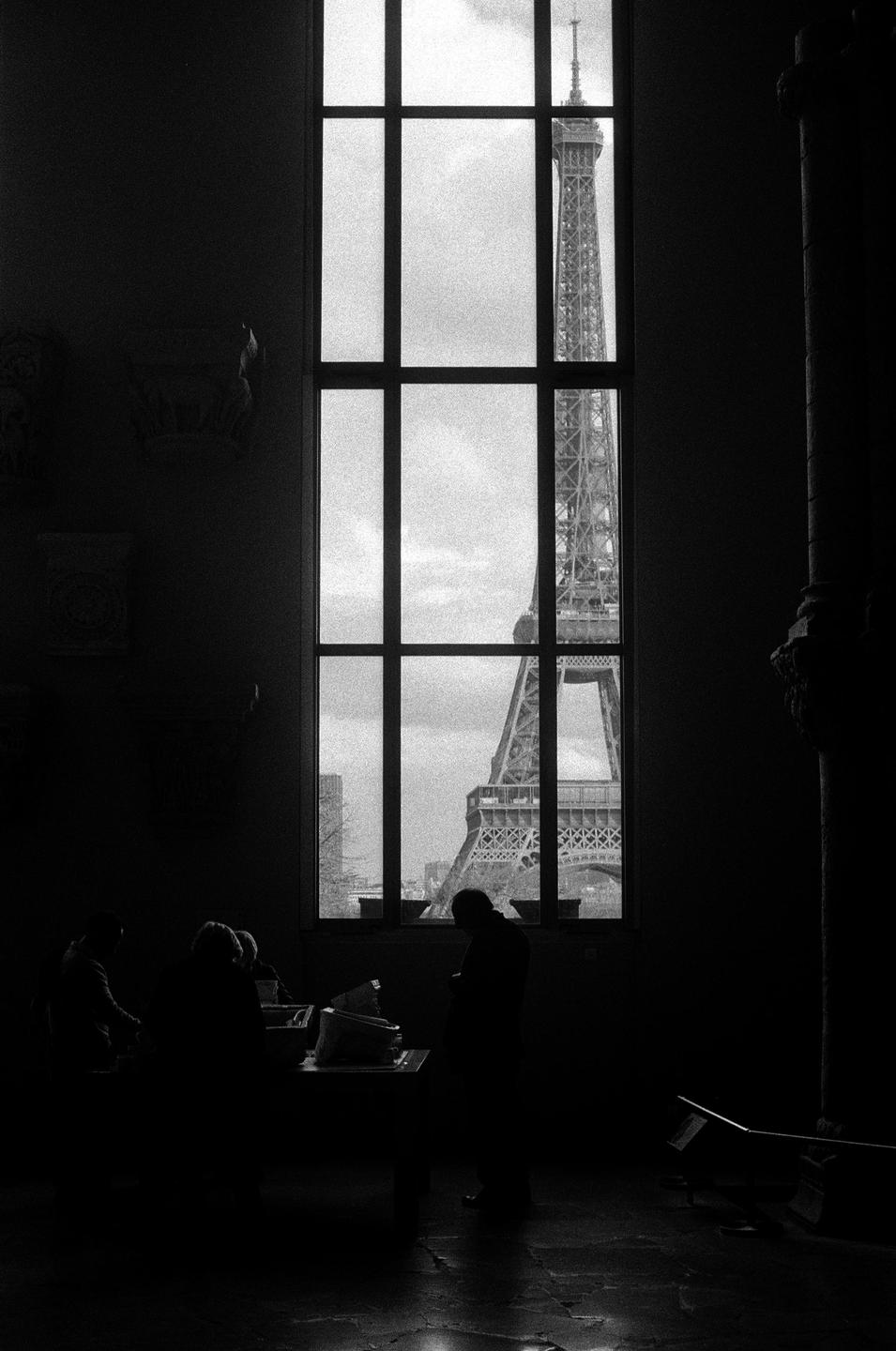 The Eiffel Tower is visible through a tall window from a dark room where people are gathered around a table.