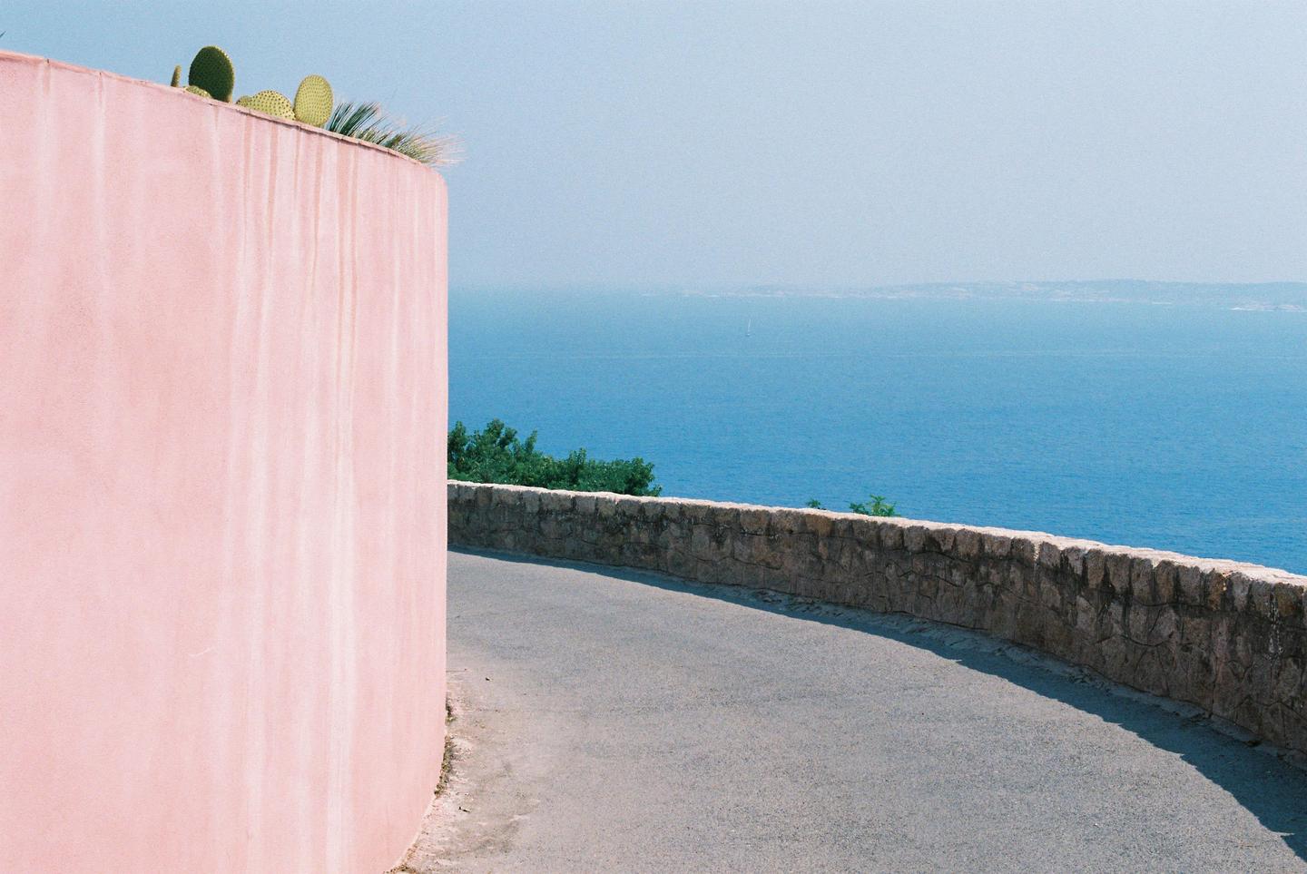 A pink wall with cacti on top stands beside a winding road overlooking a vast blue sea.