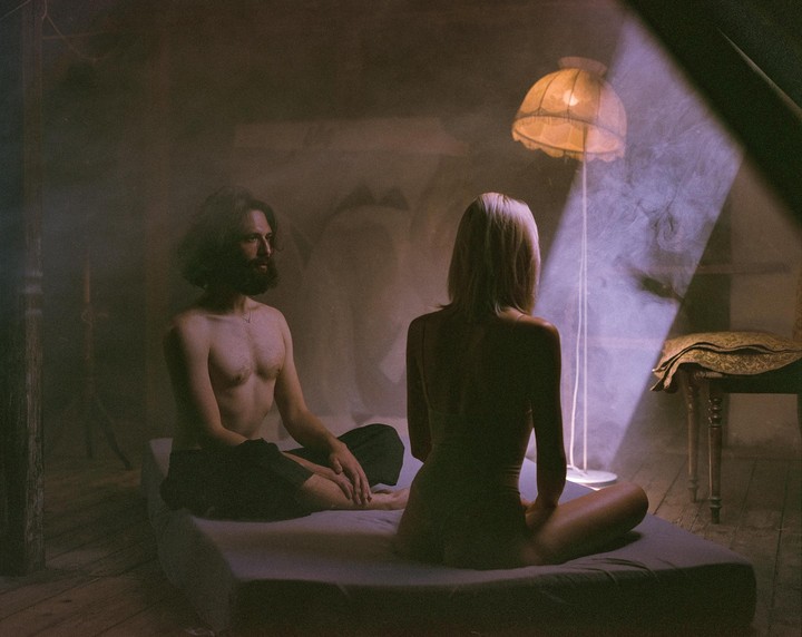 A man and a woman sit on a bed in a hazy room with a floor lamp and a beam of light.