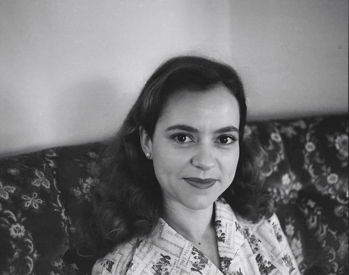 A woman with dark hair and red lipstick looks directly at the viewer while sitting on a patterned sofa.