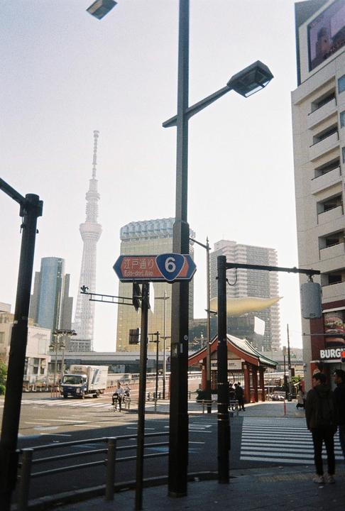 A street scene in Tokyo shows a tall tower in the distance, a street sign for Edo-dori Avenue, and people walking on the sidewalk.