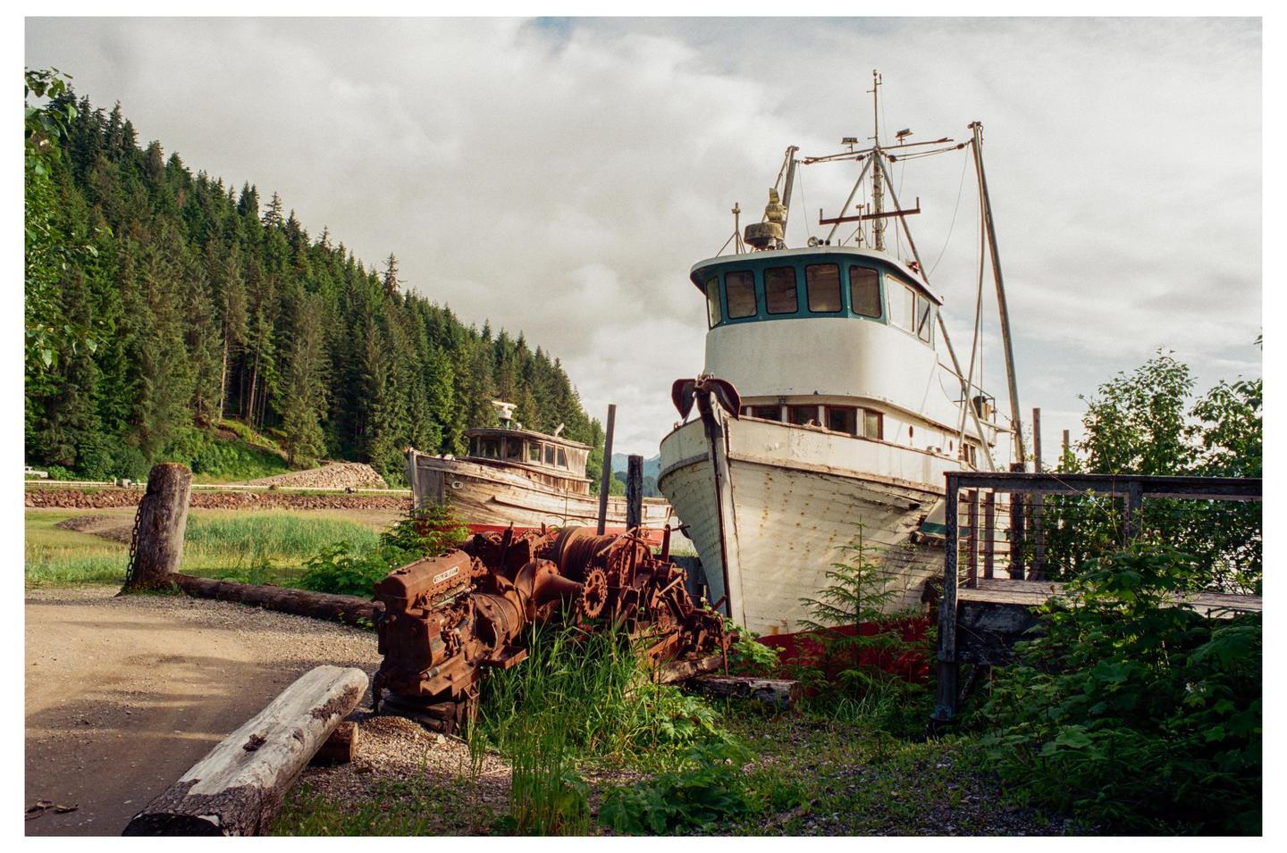 Two old fishing boats are beached on a grassy shore next to a forest under a cloudy sky.