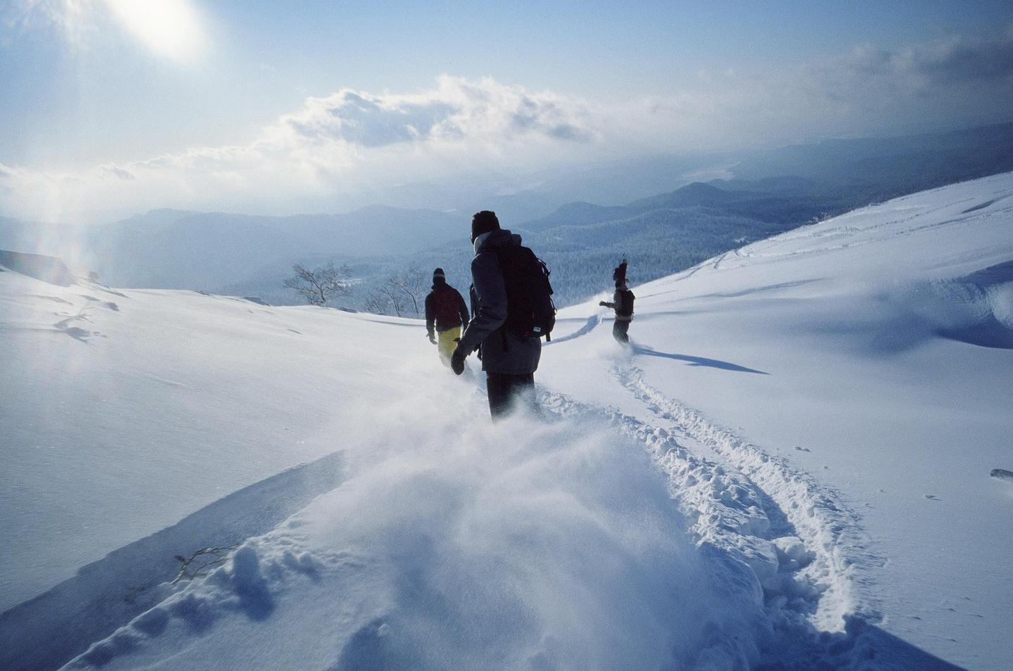 Three people walk through deep snow on a mountain with a bright sun in the sky.