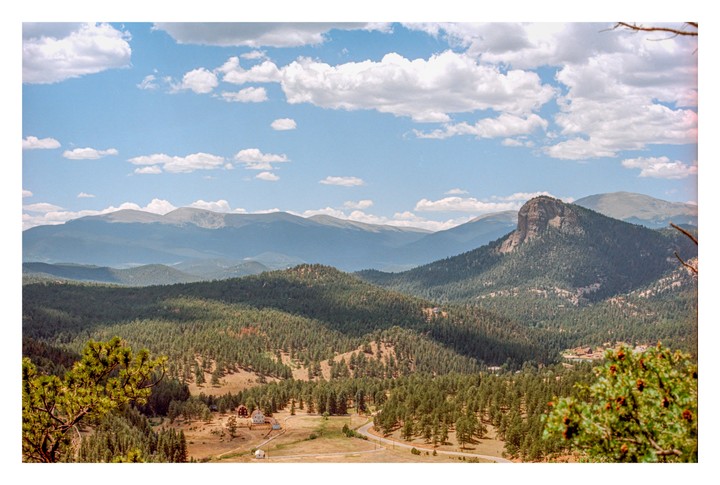 A vast landscape shows a valley with pine trees and a winding road, leading to a cluster of buildings, all set against a backdrop of mountains under a blue sky with clouds.
