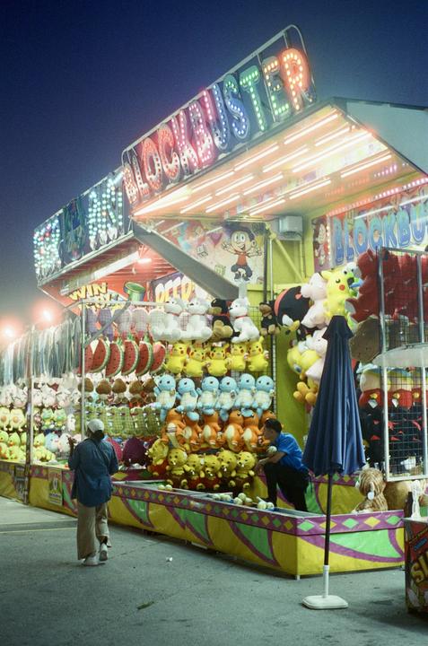 A carnival game booth with a "Blockbuster" sign is brightly lit at night, displaying numerous stuffed animal prizes.